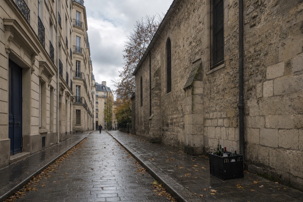 Wet narrow Paris street with limestone apartment facades, a medieval stone church wall, fallen leaves and a crate of bottles by the curb.