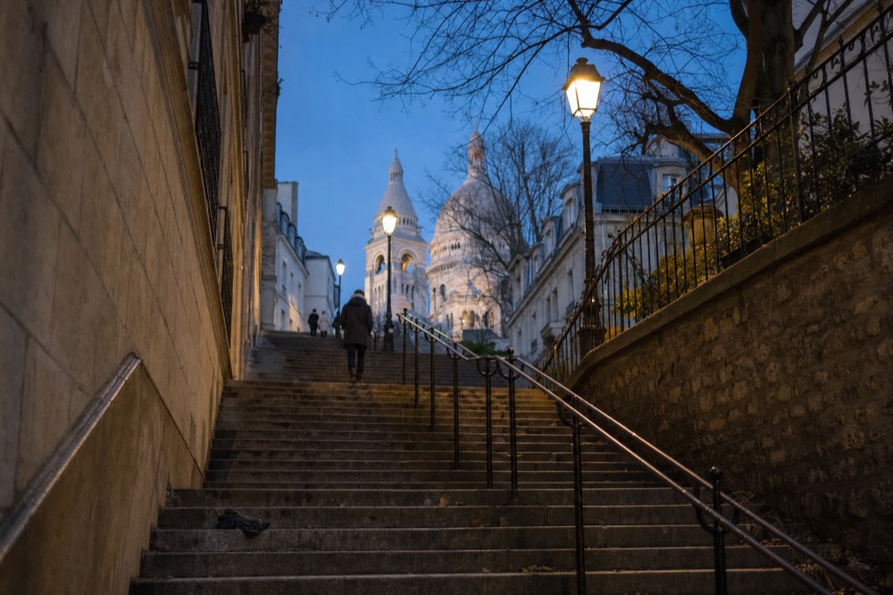 Stone stairs climb between walls and railings toward Sacré-Cœur at dusk, with lamps lit and a dropped glove on a step.
