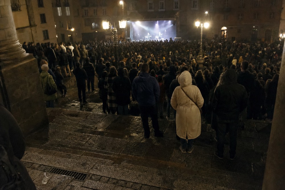 Crowd viewed from wet stone steps at night, facing a brightly lit stage in a Paris square with glowing lamp columns.
