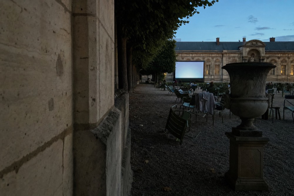 Green deckchairs face a glowing outdoor cinema screen at dusk beside clipped trees, a stone wall, and a large urn in a Paris garden.