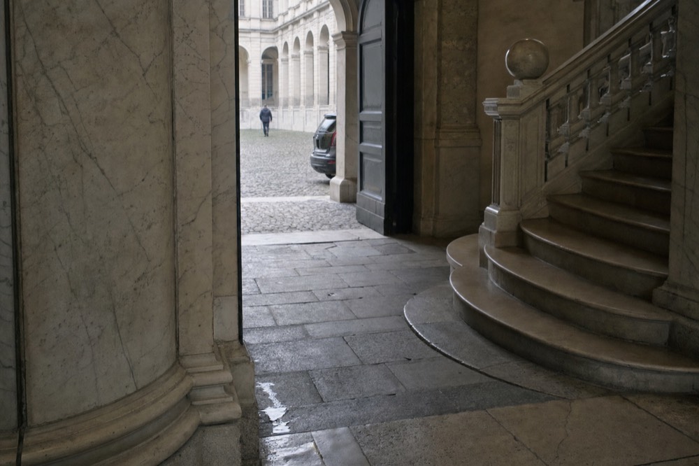 Marble entrance hall with curved stairs, an open doorway to a cobbled courtyard, a distant person, and a parked car outside.