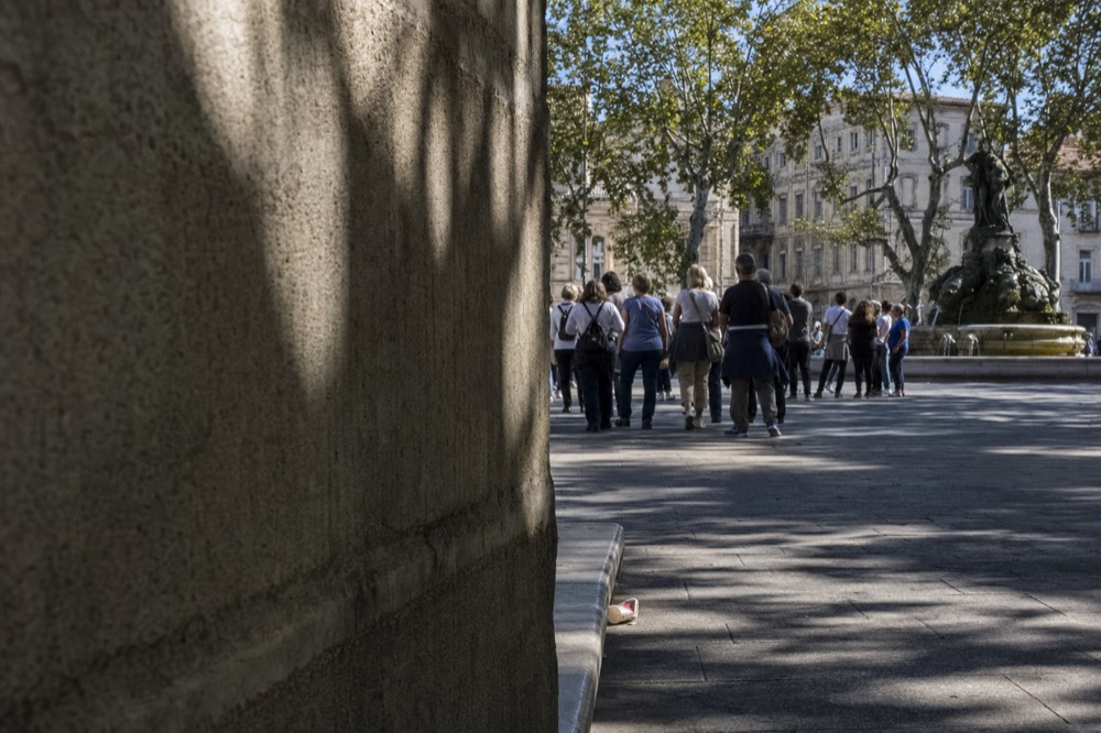 A tour group walks across a shaded Paris square past a stone wall, with a bronze fountain and plane trees in the background.