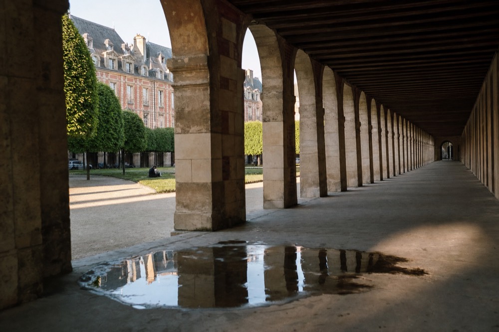 Long stone arcade beside a rose brick Paris courtyard with clipped trees, gravel, a puddle reflection, and one distant seated figure.