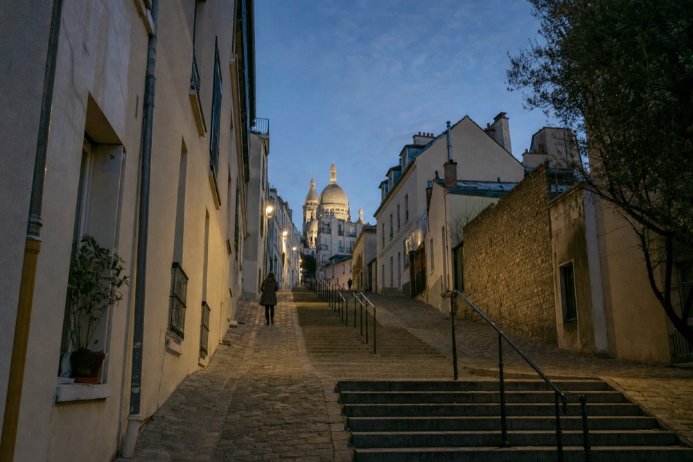 Stone steps climb between cream buildings toward the illuminated Sacre-Coeur dome at dusk, with one distant person on the path.