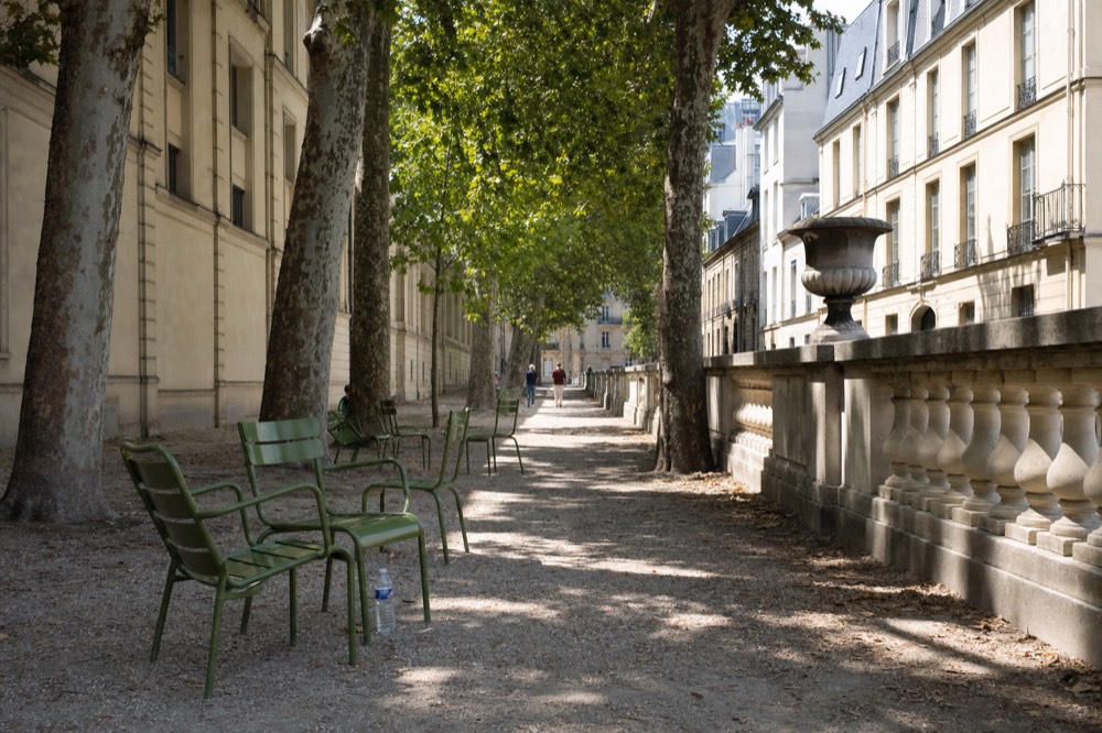 Green chairs sit on a shaded gravel alley lined with plane trees, limestone buildings, a balustrade, and a stone urn.