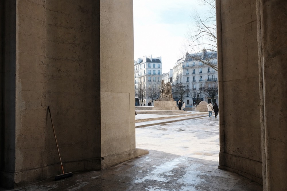 Stone archway frames a wet Parisian courtyard with a sculpture, limestone buildings, bare trees, distant pedestrians, and a broom by the wall.