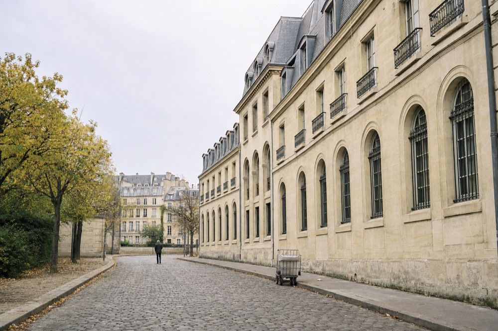 A limestone facade with arched barred windows lines a cobbled Paris street with autumn trees and a small delivery trolley.