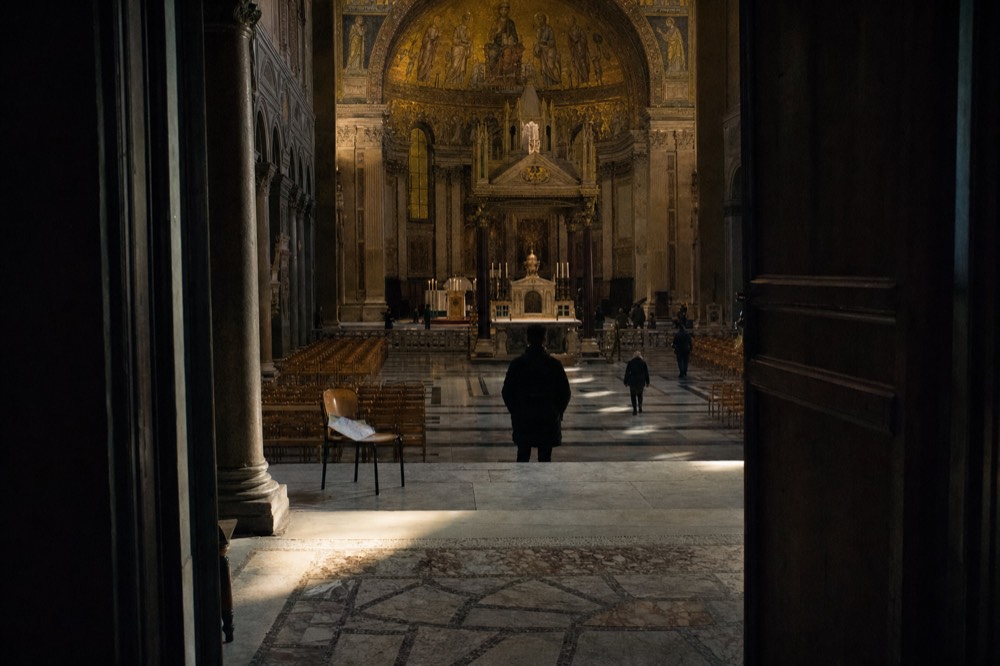 Doorway view into a dim basilica nave with sunlight on stone floors, rows of chairs, an altar, and a gold mosaic apse.