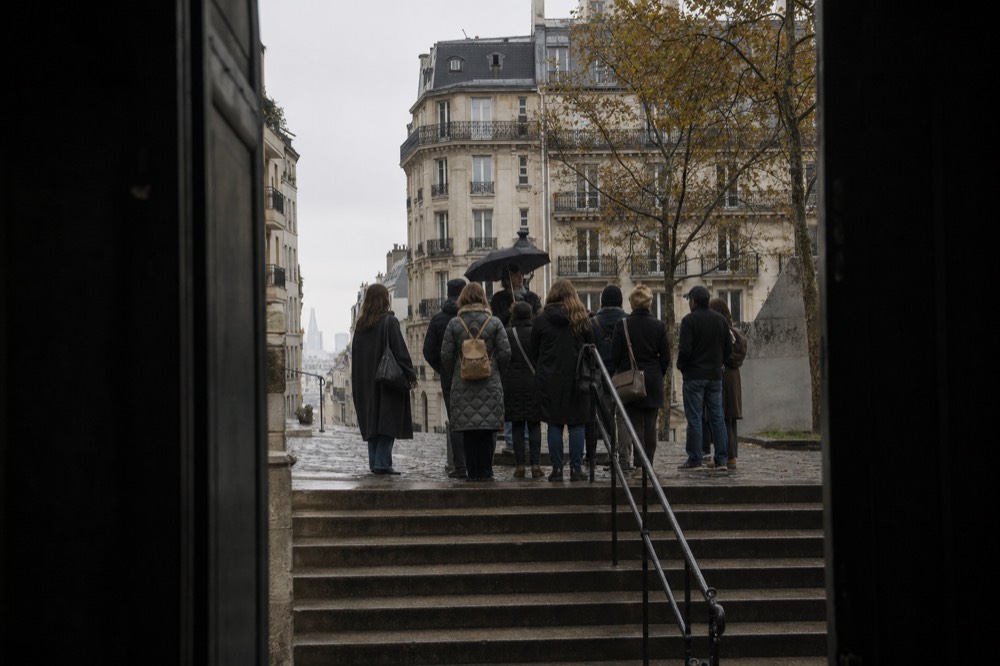 A guide with a black umbrella speaks to a small group at the top of wet stone steps between Paris apartment buildings.