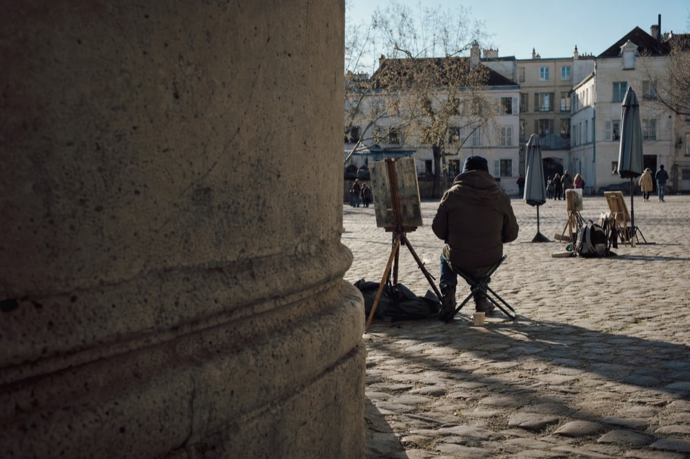 A painter sits by easels and closed umbrellas on a cobbled Paris square, seen past a stone column in low morning light.