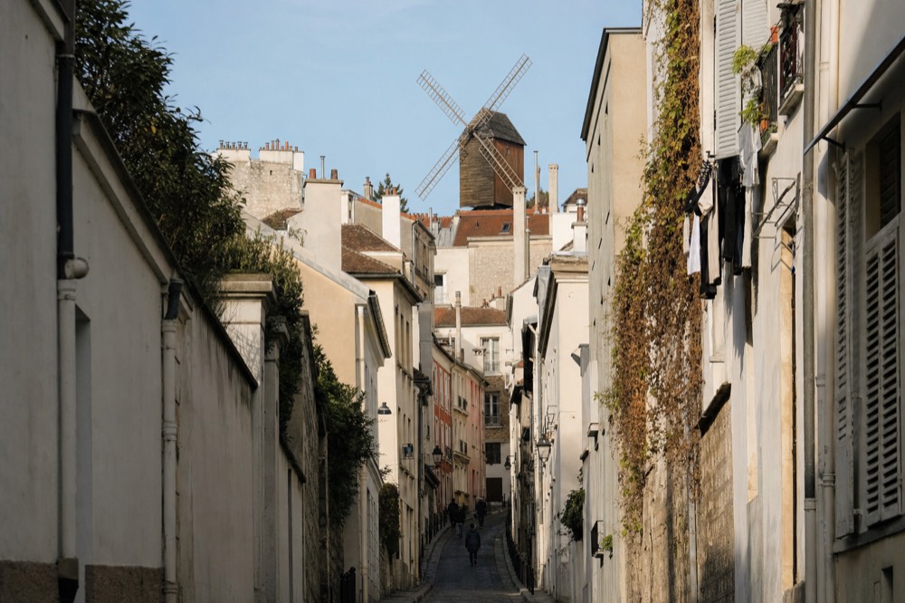 A narrow Montmartre street climbs between pale buildings toward a windmill rising above rooftops, with ivy and hanging laundry on the right.