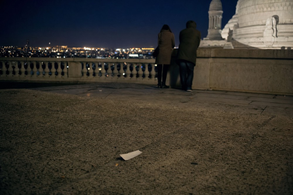 Two figures lean on a stone balustrade beside Sacre-Coeur at night, with Paris lights beyond and a paper scrap on the ground.