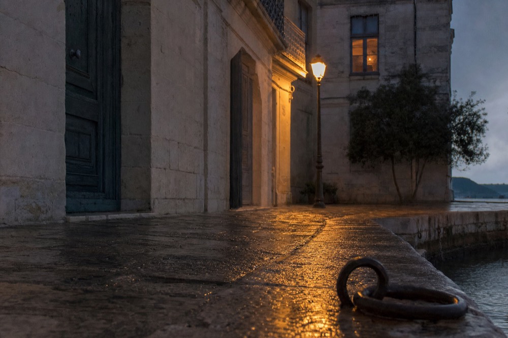 Wet cobbles on a quiet stone riverside street glow under a lamppost beside limestone walls and an iron mooring ring at night.