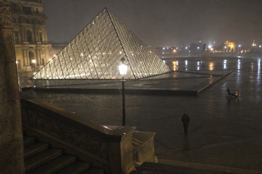 Illuminated Louvre glass pyramid stands in a wet, nearly empty courtyard at night with stone steps and a lone distant figure.
