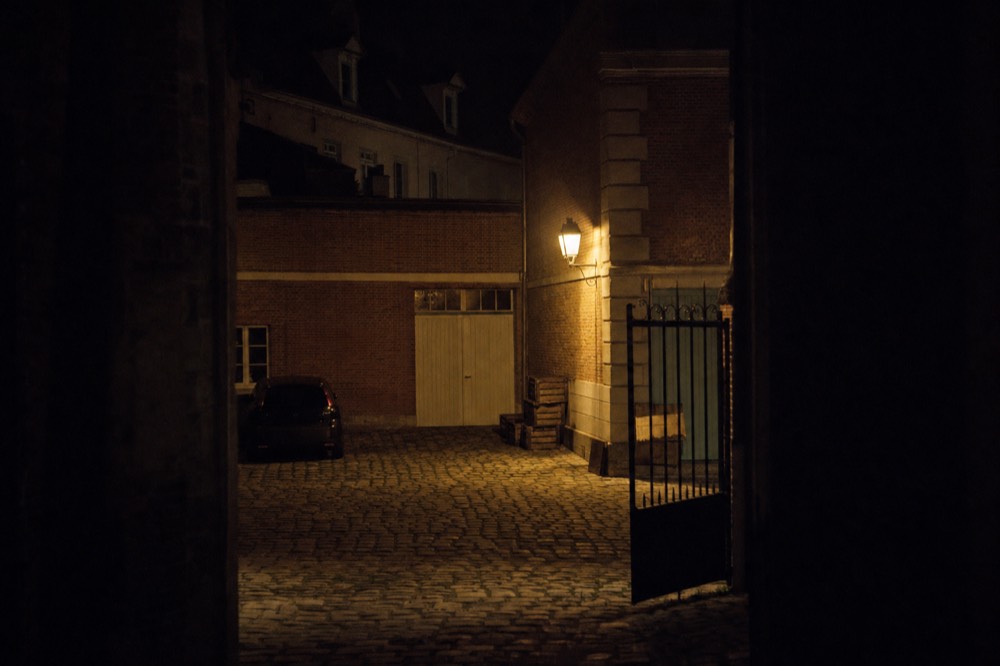 A lantern lights a cobbled brick courtyard at night, with stacked crates, an open wrought-iron gate, and a parked car in shadow.