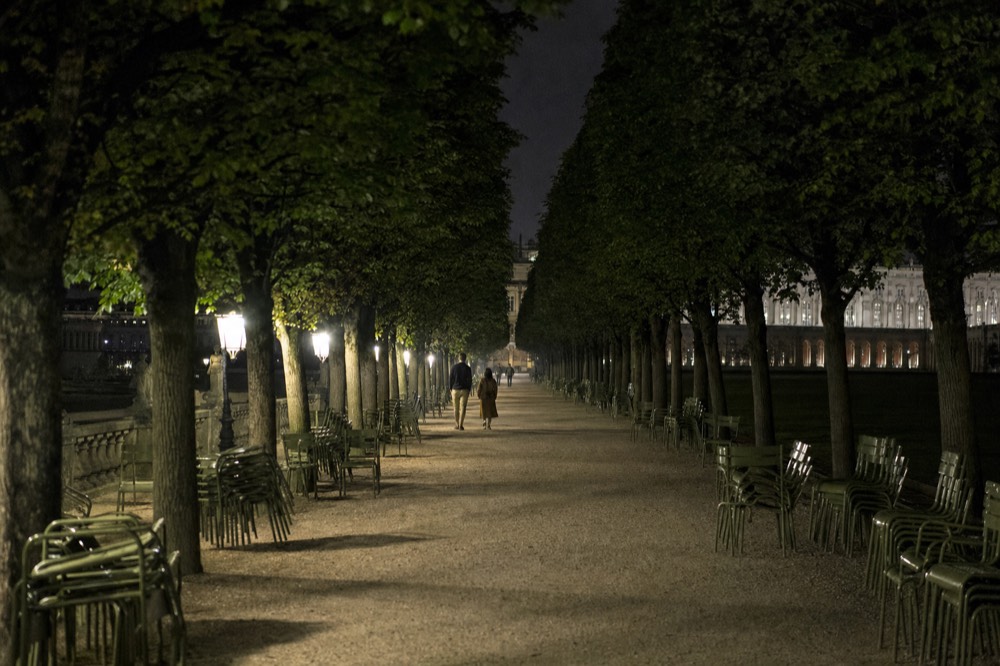Lamplit gravel park path lined with clipped trees and stacked green chairs, with two distant walkers and a glowing palace facade beyond.