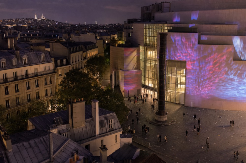 Night view over Paris rooftops to a mostly empty square where colored light projections cover a modern facade beside a tall column.