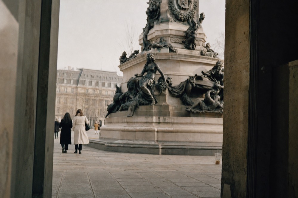 A monumental stone fountain with bronze figures stands beyond a shadowed archway as two people walk across broad paving.