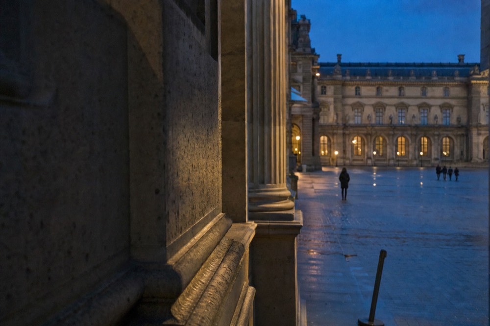 Stone columns frame the Louvre courtyard at dusk with warm lights and a few distant visitors crossing the wet paving.