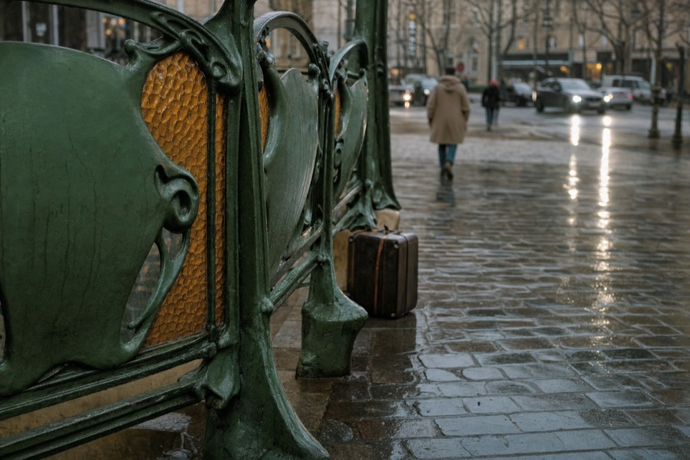 Rain-slick Paris metro entrance railing with amber glass panels, a suitcase on cobbles, and pedestrians crossing a wet street.