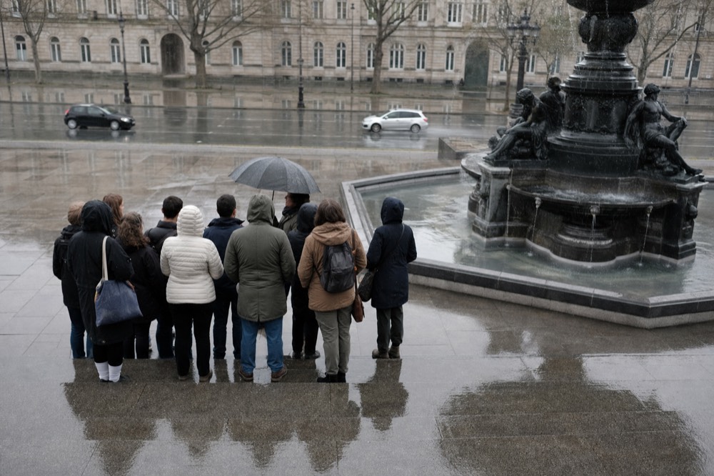 A small group stands with a guide under an umbrella beside a dark stone fountain on a wet Paris square.