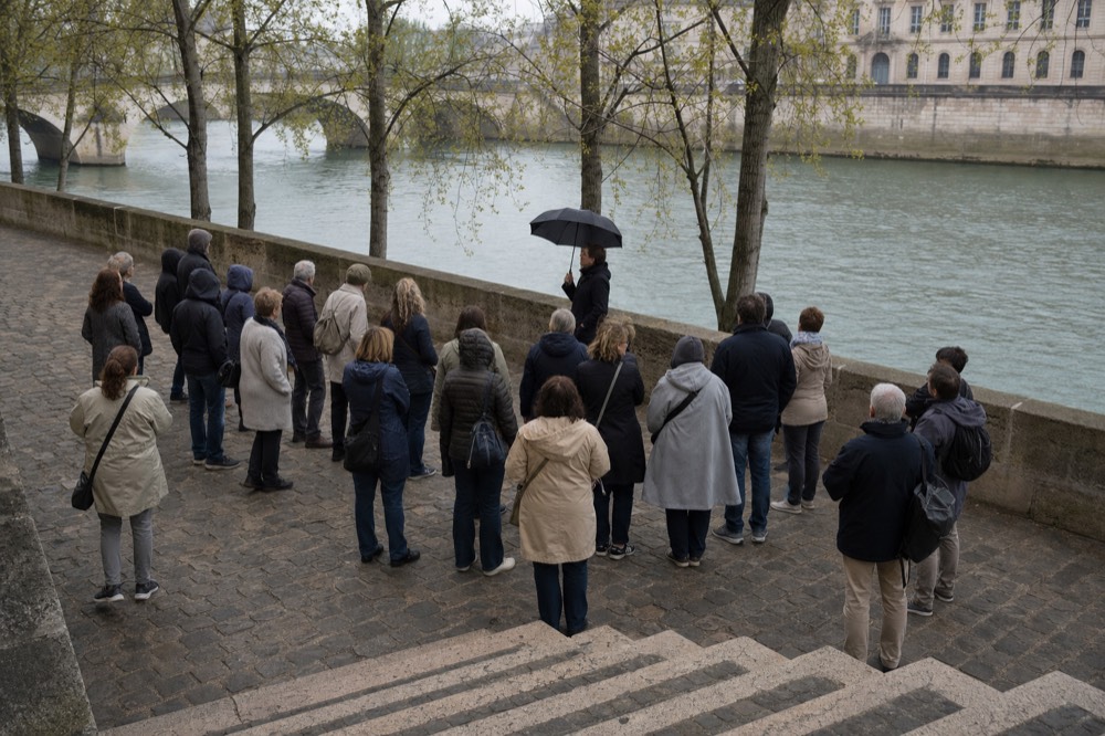 A guide with a black umbrella addresses a walking tour group on a cobbled Seine quay under spring trees.