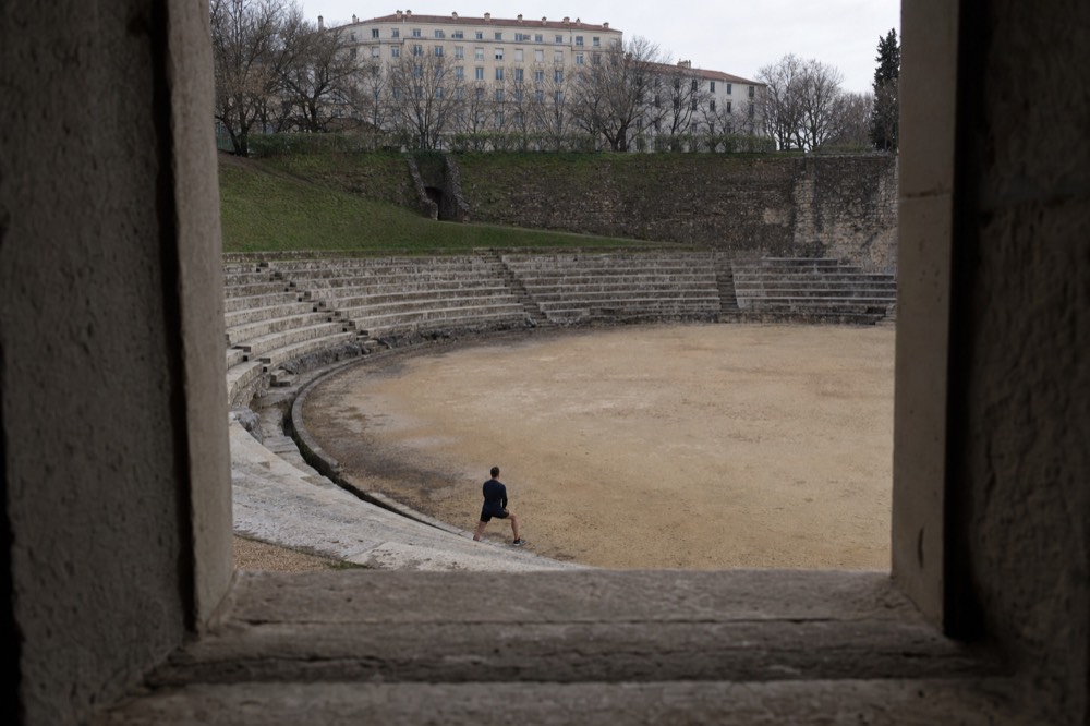 Stone amphitheatre tiers and a sandy arena are seen through a dark opening, with one distant visitor and apartment blocks beyond.