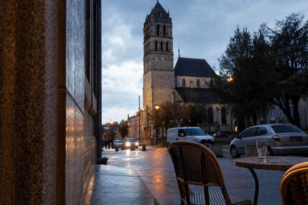Romanesque church bell tower rises beyond wet pavement, parked cars, trees, and cafe chairs with a glass on a table.