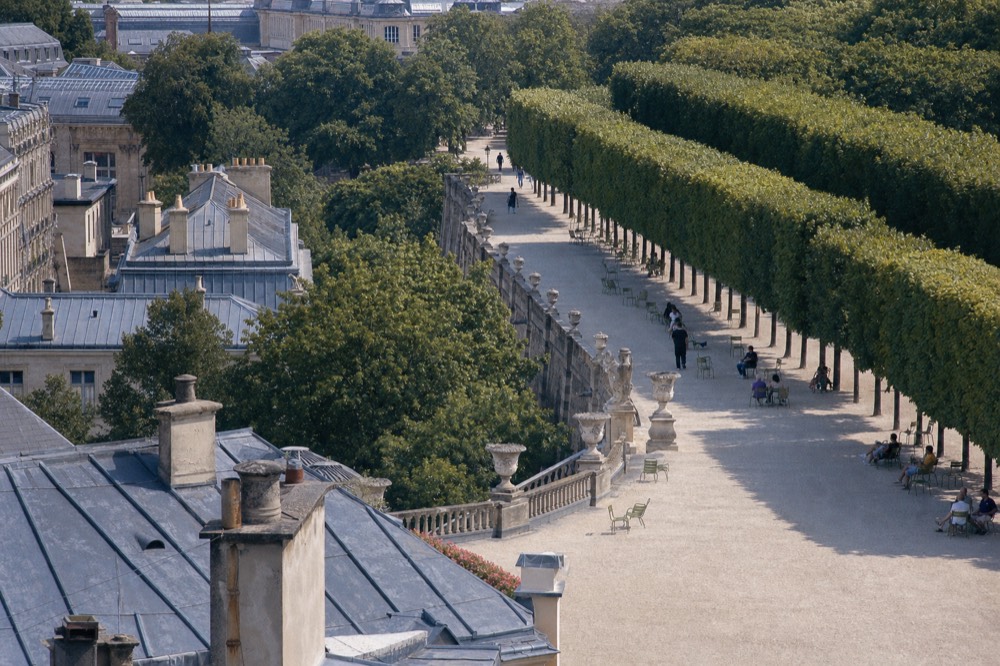 Elevated view over Paris rooftops to a gravel garden alley lined with clipped trees, green chairs, urns, and distant walkers.