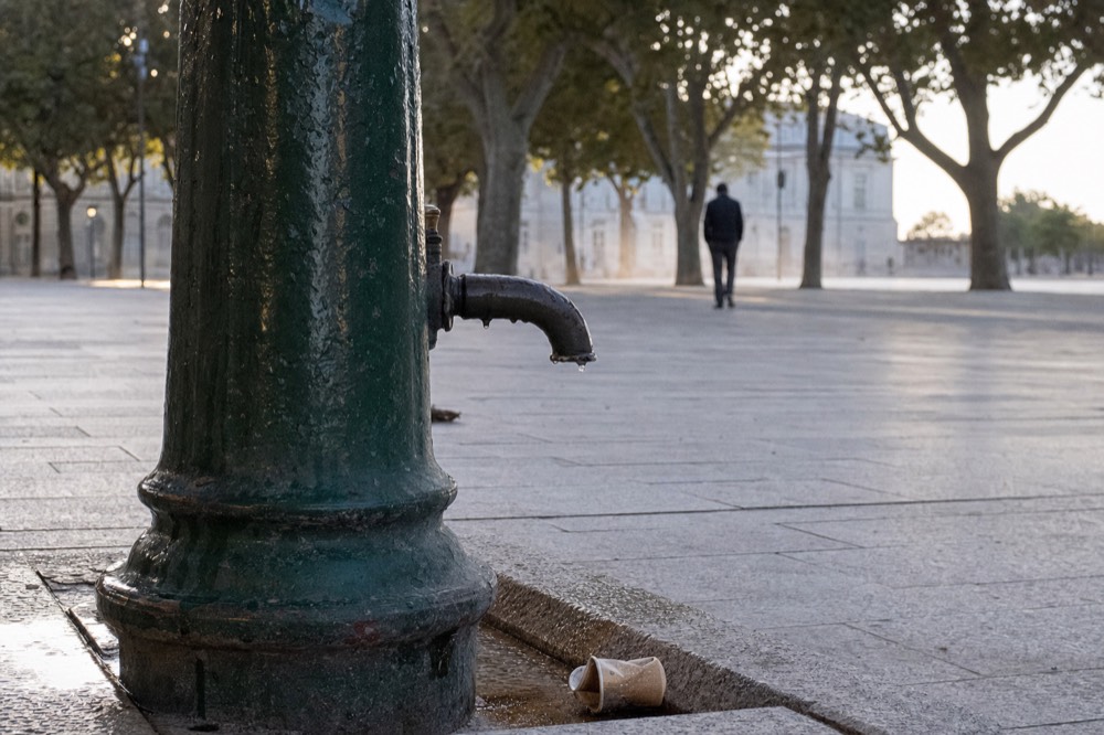 Close view of a dark green drinking fountain spout dripping onto a stone pavement with trees and a distant passer-by behind.