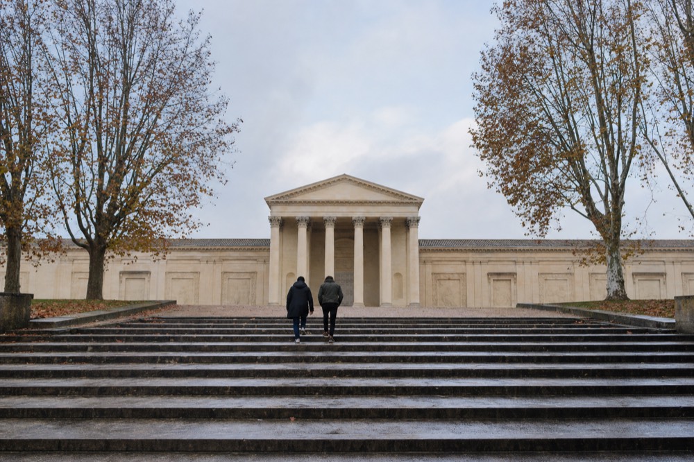 Two people climb wet stone steps between bare plane trees toward a pale neoclassical portico under a cloudy sky.
