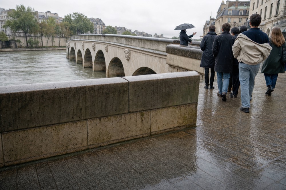 A guide holding an umbrella leads a small group along a wet stone bridge over the Seine under a grey sky.