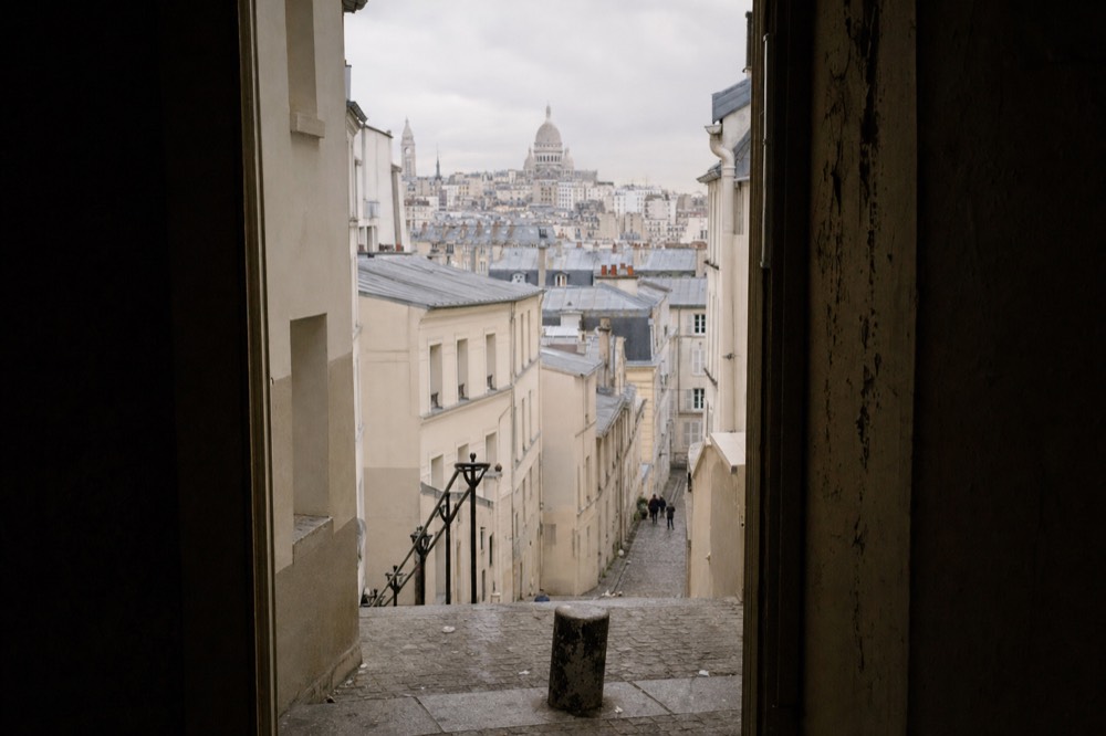 Steep Montmartre stone steps descend between cream buildings, with grey rooftops and the Sacre-Coeur dome under a cloudy sky.