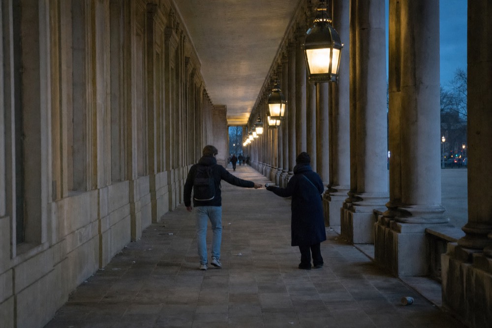 Two people exchange a small item beneath a long lit stone arcade at dusk, with columns, paving and distant walkers.