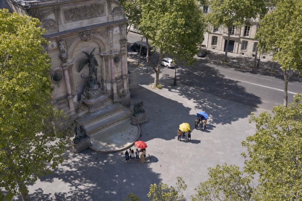 Small groups gather under red, yellow and blue umbrellas on a paved Paris square beside a large stone fountain and plane trees.