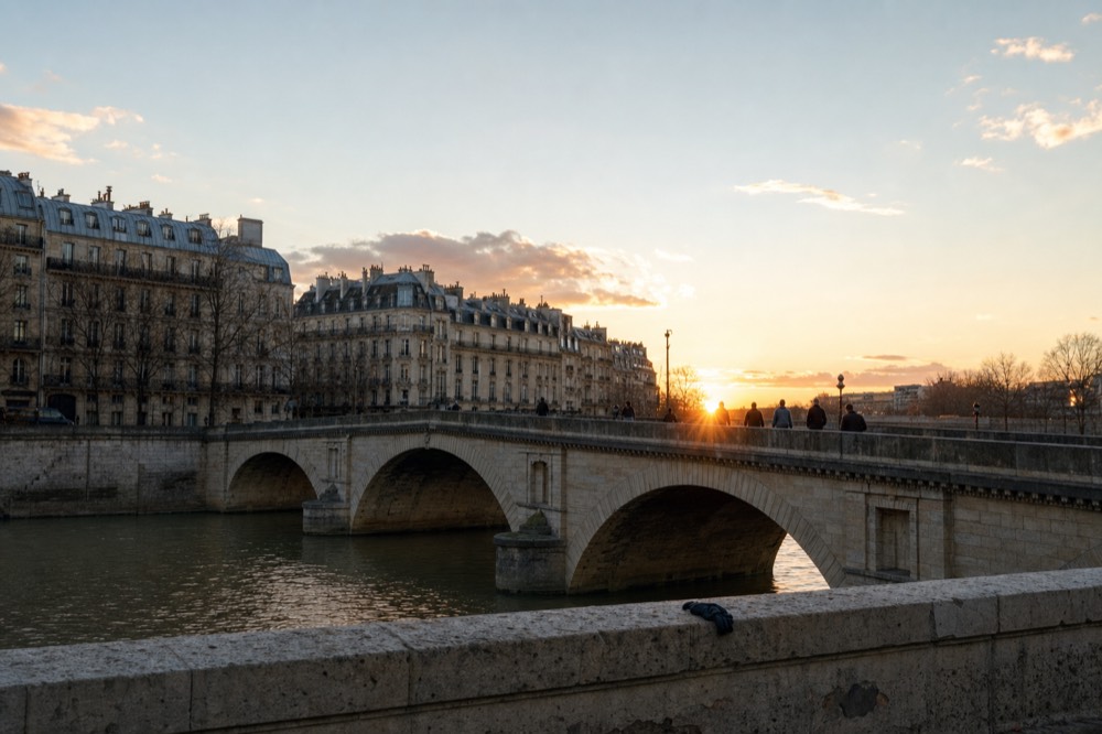 Small figures cross a stone Paris bridge over the Seine at sunset, with pale apartment blocks and a dropped glove on the parapet.
