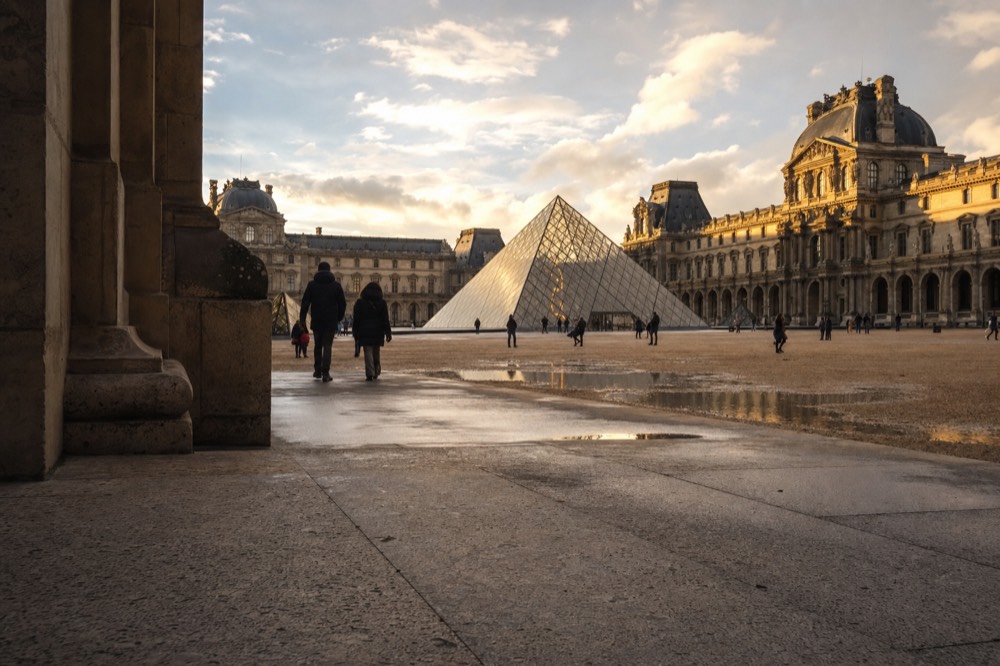 Glass Louvre Pyramid in the Cour Napoleon at golden hour, with wet paving, puddles, limestone palace facades, and distant people.