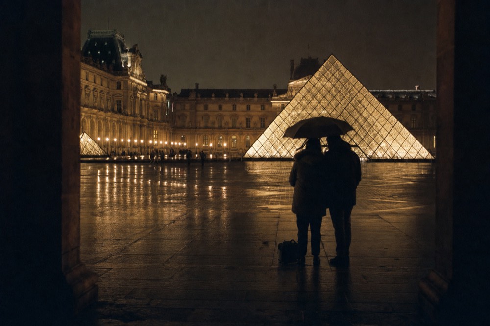 Two silhouetted people share an umbrella on wet paving before the glowing Louvre Pyramid and floodlit palace at night.