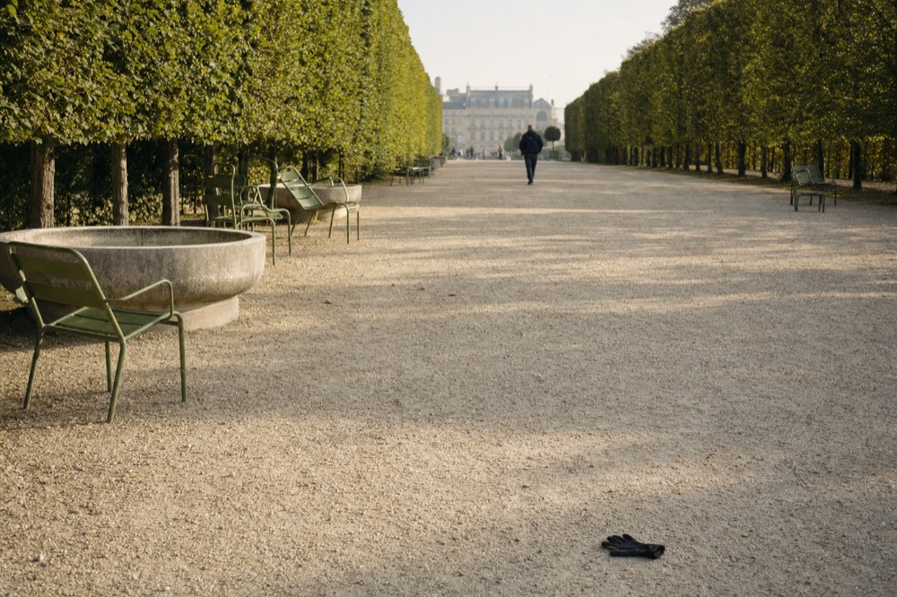 A gravel alley in the Tuileries Garden with green chairs, stone basins, clipped trees, a distant palace facade and a glove on the ground.