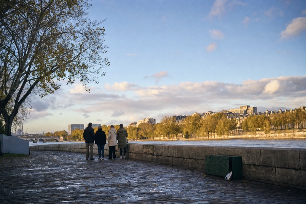 A small group stands on a wet stone quay beside the Seine, with plane trees and Parisian buildings across the river.