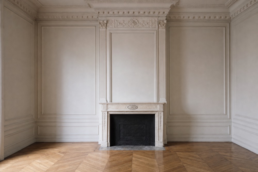 Empty ornate room with pale wall paneling, parquet floor, and a carved fireplace centered on the back wall.