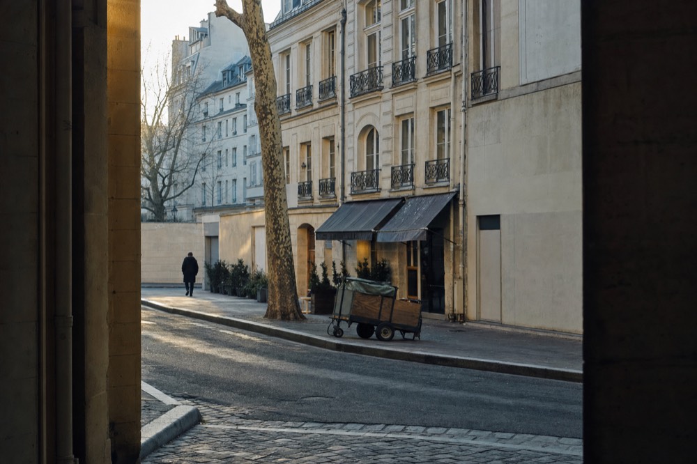 Narrow Paris street with stone facades, wrought-iron balconies, a plane tree, a delivery cart and a distant pedestrian.