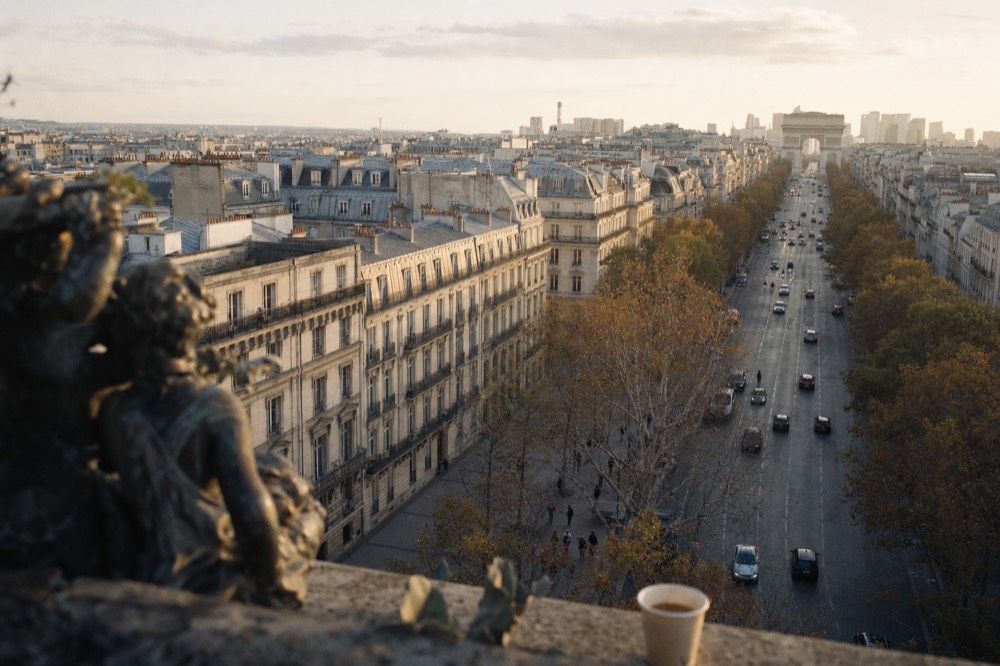 Elevated view over a Paris boulevard lined with Haussmann buildings, autumn trees, traffic, a blurred statue and a paper cup.
