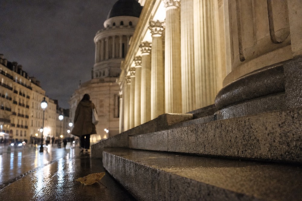 Wet granite steps and lit limestone columns lead toward a domed Paris monument at night, with a person walking away and a leaf on the pavement.