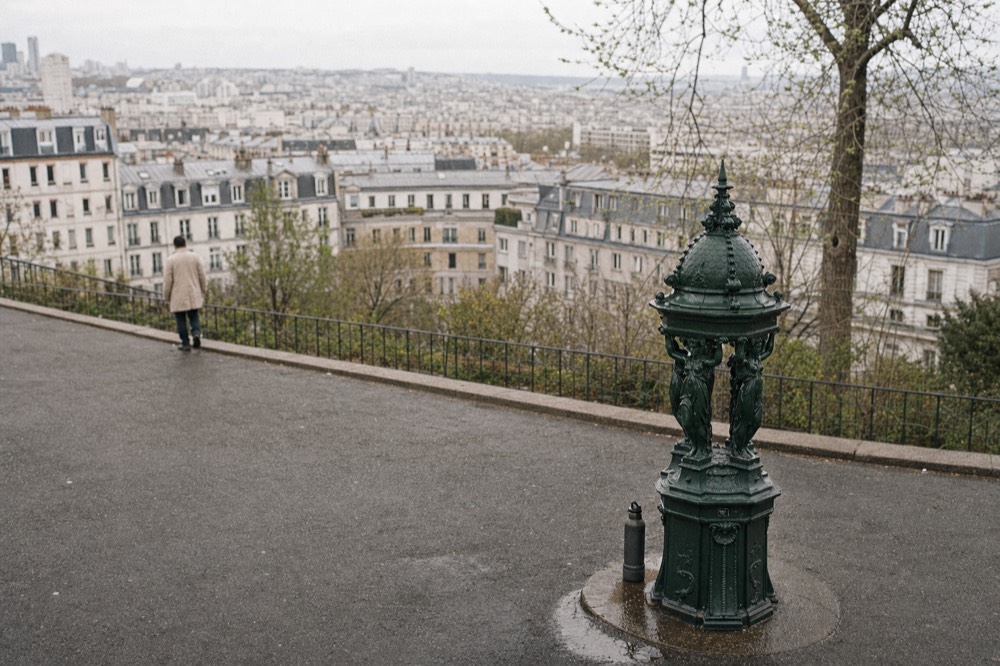 A green Wallace fountain stands on a wet Paris overlook pavement with zinc-roofed apartment blocks and a distant pedestrian behind it.
