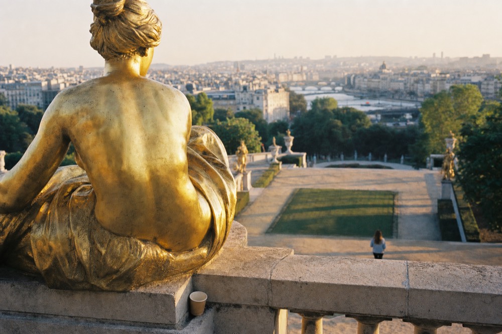 Gilded statue on a stone terrace overlooks Paris, the Seine, garden paths, a paper cup and a distant figure at golden hour.