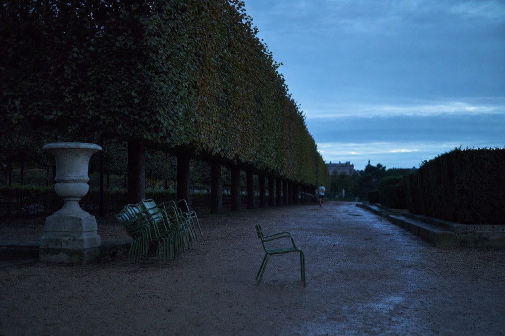 Dusk in a Paris park with clipped tree rows, wet gravel, stacked green chairs, a stone urn, and a distant jogger.