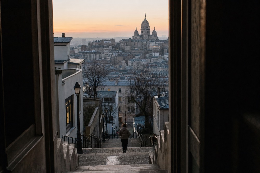 Dark doorway frames wet Montmartre steps with a lone figure, grey rooftops, bare trees and Sacre-Coeur at dawn.