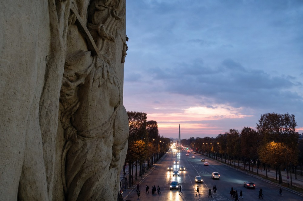 Stone relief on the Arc de Triomphe frames the Champs-Elysees at dusk, with traffic lights, trees, and the Concorde obelisk in the distance.