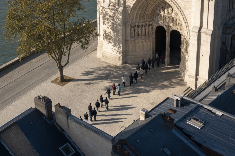 Visitors queue across the stone parvis beside a plane tree at a carved limestone portal of Notre-Dame, with rooftops and the river nearby.